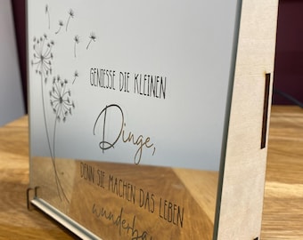 May include: A wooden frame with a glass mirror inside. The mirror has a dandelion design and the text "Geniesse die kleinen Dinge, denn sie machen das Leben wunderbar."  The frame is standing on a wooden surface.