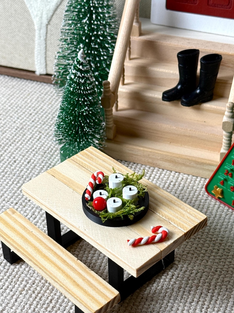 May include: Miniature Christmas scene featuring a wooden picnic table with a black base. A black tray holds four white candles, red berries, and greenery. Candy canes and miniature Christmas trees complete the festive display. Black boots sit on the porch steps.