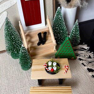May include: Miniature Christmas scene featuring a red door with a green wreath, small green trees, and a picnic table. The table has a small black bowl with white candles, a red ball, and candy canes. Black boots are on the porch.