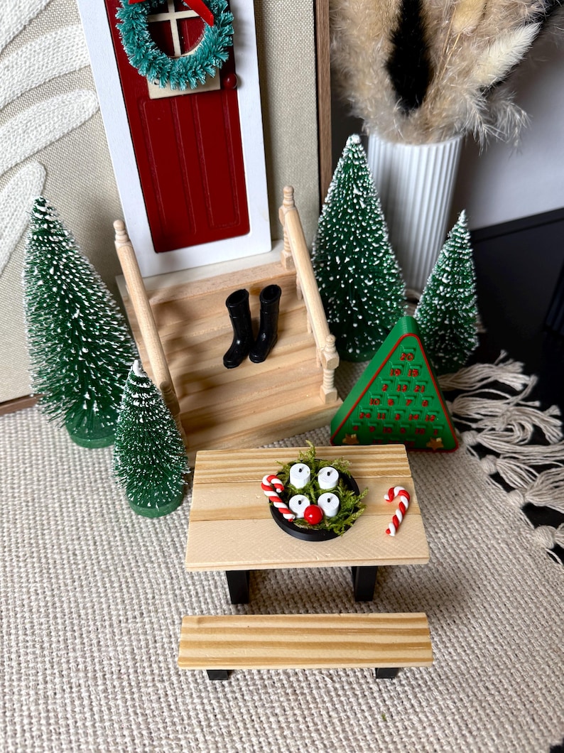 May include: Miniature Christmas scene featuring a red door with a green wreath, small green trees, and a picnic table. The table has a small black bowl with white candles, a red ball, and candy canes. Black boots are on the porch.