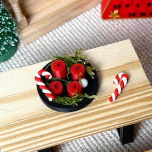 May include: Miniature Christmas scene featuring a small wooden table with a black plate holding four red candles, greenery, and two candy canes. A small green Christmas tree and a red advent calendar are in the background.