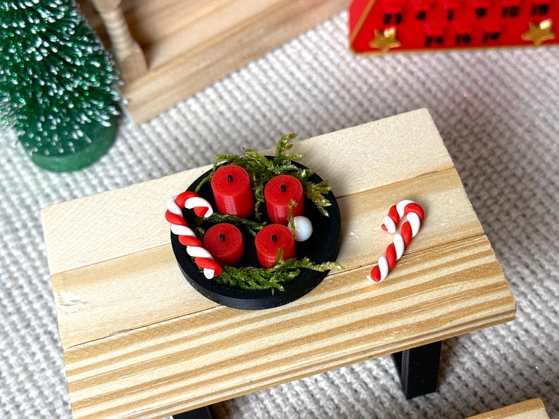 May include: Miniature Christmas scene featuring a small wooden table with a black plate holding four red candles, greenery, and two candy canes. A small green Christmas tree and a red advent calendar are in the background.