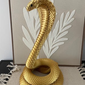 May include: A golden cobra sculpture with a detailed, textured surface. The snake is coiled with its head raised, showcasing intricate scale patterns. The sculpture is a decorative item, likely for home decor.
