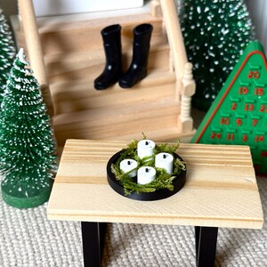 May include: Miniature Christmas scene featuring a small wooden table with a black rimmed candle holder filled with white candles and green moss. Two black boots sit on the stairs. A green Christmas tree and a green Advent calendar are also present.