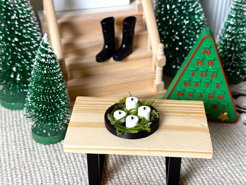 May include: Miniature Christmas scene featuring a small wooden table with a black rimmed candle holder filled with white candles and green moss. Two black boots sit on the stairs. A green Christmas tree and a green Advent calendar are also present.