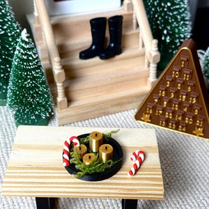 May include: Miniature Christmas scene featuring a wooden table with a black tray holding four gold candles and greenery, plus two candy canes. A brown pyramid advent calendar and black boots on a porch are also visible. Green miniature Christmas trees complete the holiday setting.