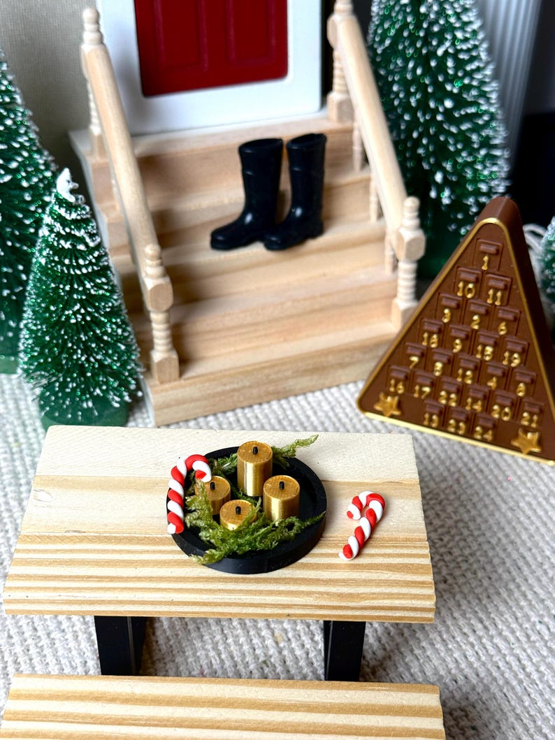 May include: Miniature Christmas scene featuring a wooden table with a black tray holding four gold candles and greenery, plus two candy canes. A brown pyramid advent calendar and black boots on a porch are also visible. Green miniature Christmas trees complete the holiday setting.