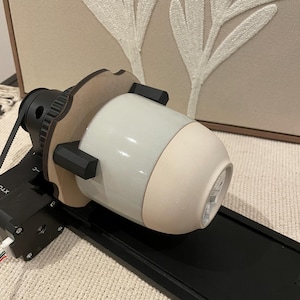 May include: A ceramic vase with a light blue and beige color scheme is mounted on a black machine. The machine has a wooden frame and black supports. The background features a beige canvas with white embroidered leaf designs.