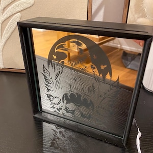 Puede incluir: Un espejo con marco negro que tiene un panel de vidrio con un diseño grabado de un cuervo, una calavera y elementos florales. El cuervo está posado en una rama con las alas extendidas. La calavera está rodeada de flores y hojas. El fondo es de color marrón oscuro.