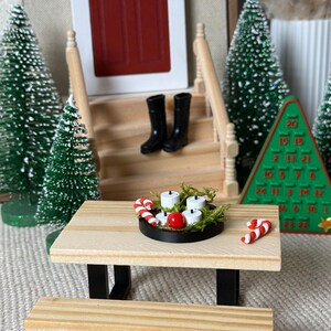 May include: Miniature Christmas scene with a wooden picnic table, bench, and a black tray holding candles, a red ball, and candy canes. Black boots sit on the porch steps. Green Christmas trees and a Christmas tree-shaped advent calendar complete the festive display.