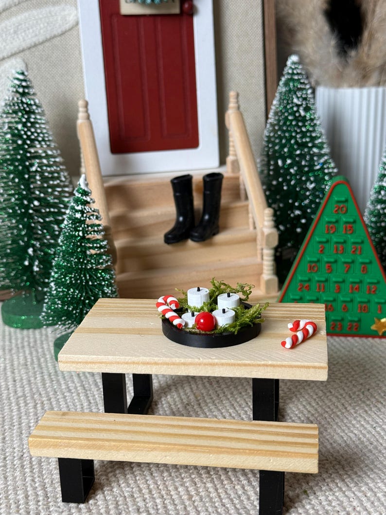 May include: Miniature Christmas scene with a wooden picnic table, bench, and a black tray holding candles, a red ball, and candy canes. Black boots sit on the porch steps. Green Christmas trees and a Christmas tree-shaped advent calendar complete the festive display.