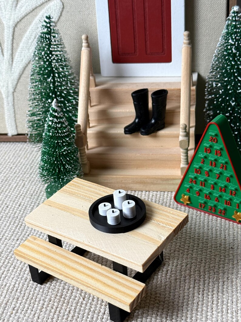 May include: Miniature Christmas scene with a wooden picnic table, black boots on steps, and a green advent calendar. The table holds a black tray with four white candles. Decorative miniature Christmas trees are also present.