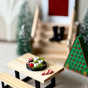 May include: Miniature Christmas scene featuring a wooden picnic table with a black base. A small black tray holds four white candles, red berries, and greenery. Two candy canes and a green Christmas tree are also visible.