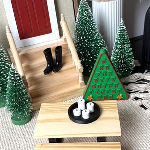 May include: Miniature Christmas scene with a red door, wooden stairs, and small black boots. A green tree-shaped advent calendar and several miniature Christmas trees are arranged around a small wooden table with a black tray holding white candles.