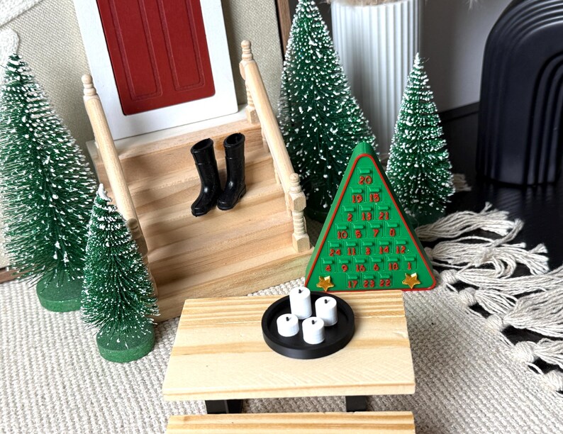 May include: Miniature Christmas scene with a red door, wooden stairs, and small black boots. A green tree-shaped advent calendar and several miniature Christmas trees are arranged around a small wooden table with a black tray holding white candles.