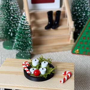 May include: Miniature Christmas scene with a small wooden table holding a black tray with white candles, a red ball, and green moss. Two candy canes and miniature black boots are also visible.