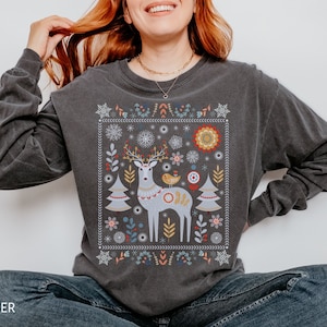 May include: A dark gray long-sleeved shirt with a colorful embroidered design of a reindeer, a bird, and a Christmas tree. The design is surrounded by a border of snowflakes and other winter motifs.