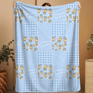 May include: A light blue patchwork blanket featuring cartoon ducklings wearing blue hats. The blanket has a gingham, dotted, and striped pattern. The name "Oliver" is printed on the blanket.