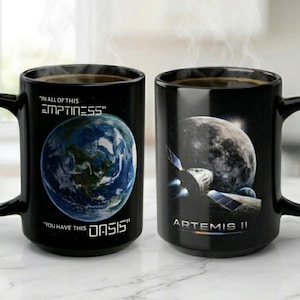 May include: Two black ceramic mugs with coffee. One mug features a blue Earth graphic with the text "IN ALL OF THIS EMPTINESS, YOU HAVE THIS OASIS." The other mug has a moon and spacecraft graphic with "ARTEMIS II."