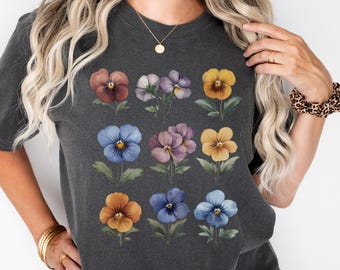 May include: A dark gray t-shirt with a colorful floral print of pansies. The pansies are arranged in three rows of four, with each row featuring a different color combination. The shirt has a relaxed fit and a crew neck.