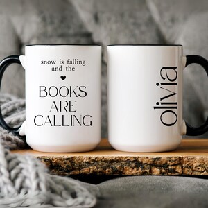 May include: Two white ceramic mugs with black handles and rims. One mug has the text "snow is falling and the BOOKS ARE CALLING" and a heart symbol. The other mug has the name "Olivia" in a black font.