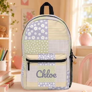 May include: A backpack featuring a patchwork design of lavender, light green, and cream panels with floral, polka dot, and striped patterns. The front pocket displays the name Chloe in a purple script font and includes a black zipper closure.