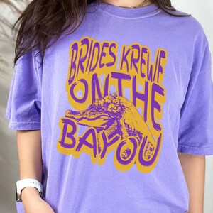 May include: Purple t-shirt with a gold graphic of an alligator wearing a bridal veil and the text "Brides Krewe On The Bayou".