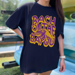 May include: A navy blue t-shirt with a graphic design that reads "BACH ON THE BAYOU" in purple and yellow. The design features an alligator. The shirt is a casual, oversized fit.