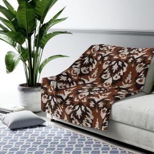 May include: A brown, black, and white patterned throw blanket draped over a white sofa. The blanket features a repeating leaf-like design. A large potted plant is to the left of the sofa. A blue patterned rug is on the floor.