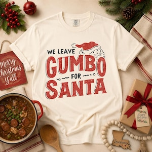 May include: A cream-colored short-sleeve t-shirt featuring a red and black graphic print that reads WE LEAVE GUMBO FOR SANTA, accompanied by a Santa hat illustration. The shirt has a crew neckline and a distressed, vintage-style ink texture.