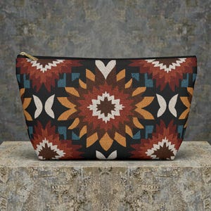 May include: A rectangular cosmetic bag with a black background and a geometric pattern in shades of brown, orange, white, and blue. The bag has a gold zipper and is likely made of fabric.