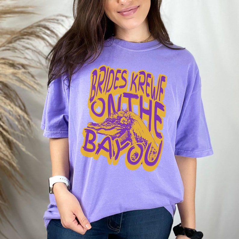 May include: Purple t-shirt with a gold graphic of an alligator wearing a bridal veil and the text "Brides Krewe On The Bayou".