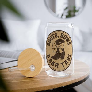 May include: A clear glass can-shaped drinking glass with a wooden lid and straw. The glass features a brown oval label with the words "BOOTS, BOWS & BAR TABS" surrounding a brown cowboy boot with a bow.