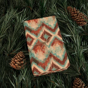 May include: A rectangular gift box wrapped in paper featuring a repeating teal and rust-colored ikat chevron pattern, tied with thin brown twine in a simple bow, resting among dark green pine needles and several brown pinecones.