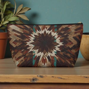 May include: A rectangular cosmetic bag with a geometric pattern in shades of brown, tan, and teal. The design features a central starburst motif. The bag has a black zipper and is likely made of fabric.