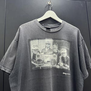 May include: Grey t-shirt with a black and white image of a group of people and a boombox. The shirt has a crew neck and short sleeves. The text "Stussy x Josh Davis" is printed on the bottom right.
