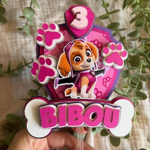 Puede incluir: Adorno para tarta rosa y blanco con un personaje de dibujos animados, el número 3 y el nombre "BIBOU". El adorno tiene forma de escudo con huellas de patas y un personaje de dibujos animados en el centro. El nombre "BIBOU" está en letras rosas.