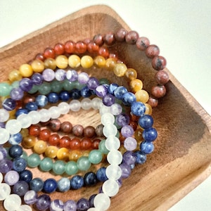 May include: A collection of beaded bracelets in various colors, including red, yellow, green, blue, and purple. The bracelets are displayed in a wooden tray, showcasing the different gemstones and their unique hues.