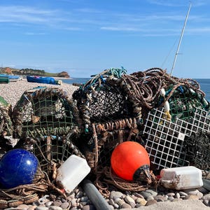 Puede incluir: Una pila de trampas para langostas y equipo de pesca en una playa de guijarros. Las trampas están hechas de malla de alambre y están apiladas unas sobre otras. También hay cuerdas, boyas y otros equipos de pesca en la pila.