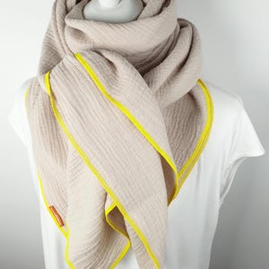 Puede incluir: Una bufanda de algod&oacute;n beige con ribete amarillo. La bufanda est&aacute; doblada y drapeada alrededor del cuello de un maniqu&iacute;.