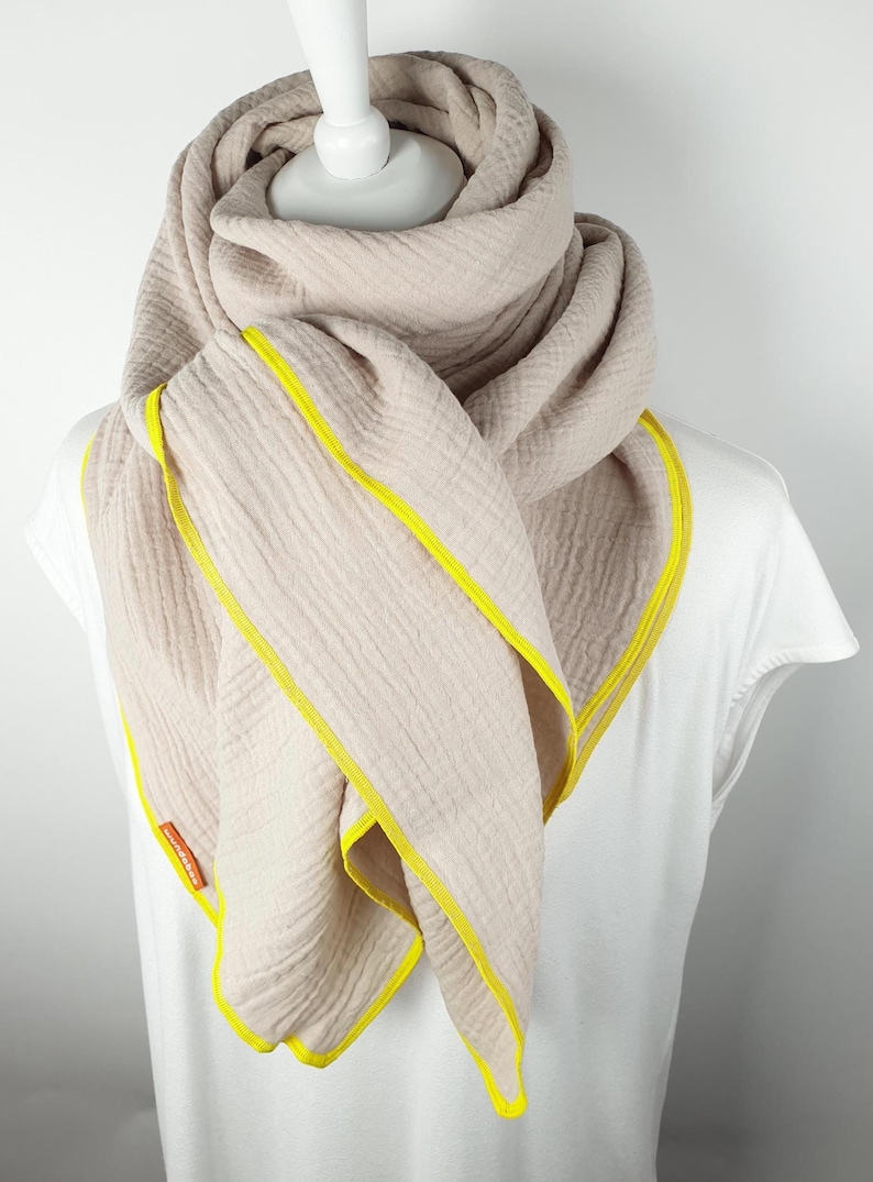 Puede incluir: Una bufanda de algod&oacute;n beige con ribete amarillo. La bufanda est&aacute; doblada y drapeada alrededor del cuello de un maniqu&iacute;.