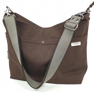 May include: A brown shoulder bag with a gray and white patterned strap. The bag has a rectangular shape with a curved top and a small pocket on the front. The strap is adjustable and attached with silver metal hardware. The brand name "wundeboo" is on a small white tag.