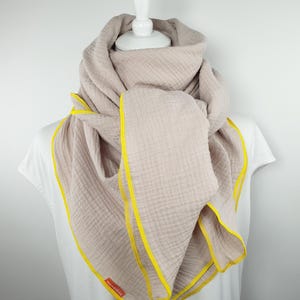 Puede incluir: Una bufanda de algod&oacute;n beige claro con un borde amarillo. La bufanda est&aacute; doblada y drapeada alrededor de un maniqu&iacute; blanco.