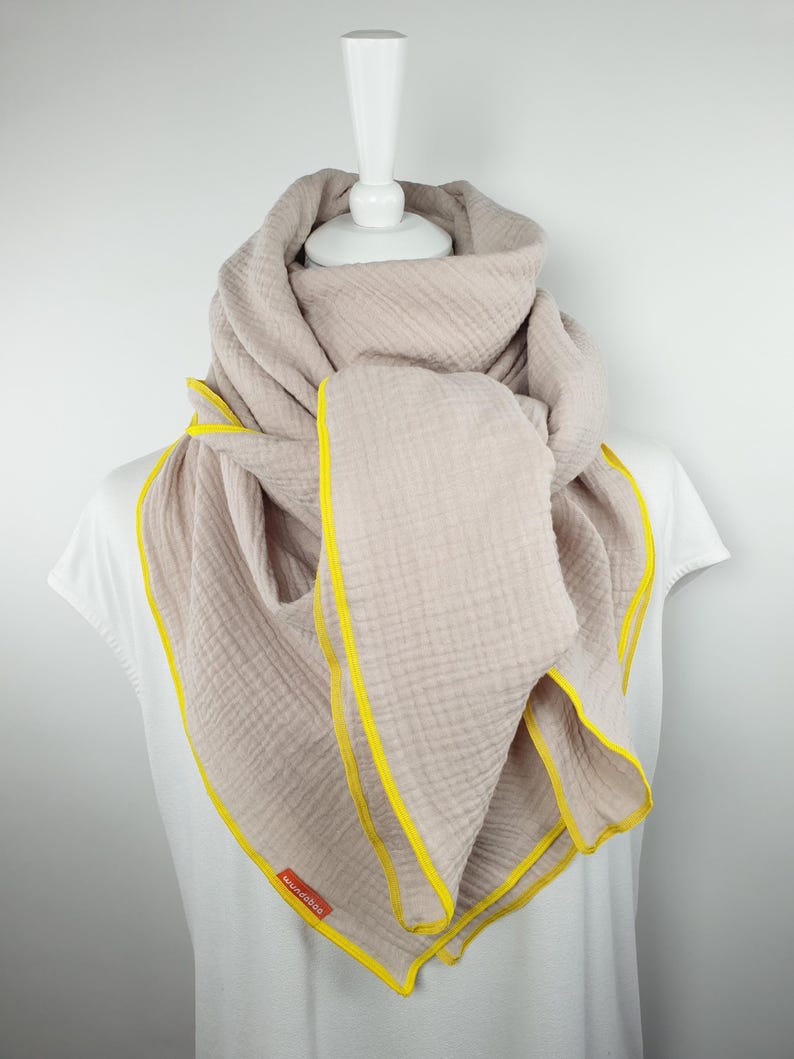 Puede incluir: Una bufanda de algod&oacute;n beige claro con un borde amarillo. La bufanda est&aacute; doblada y drapeada alrededor de un maniqu&iacute; blanco.