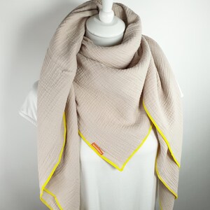 Puede incluir: Una bufanda de algod&oacute;n beige con ribete amarillo. La bufanda est&aacute; doblada y drapeada sobre un maniqu&iacute; blanco.