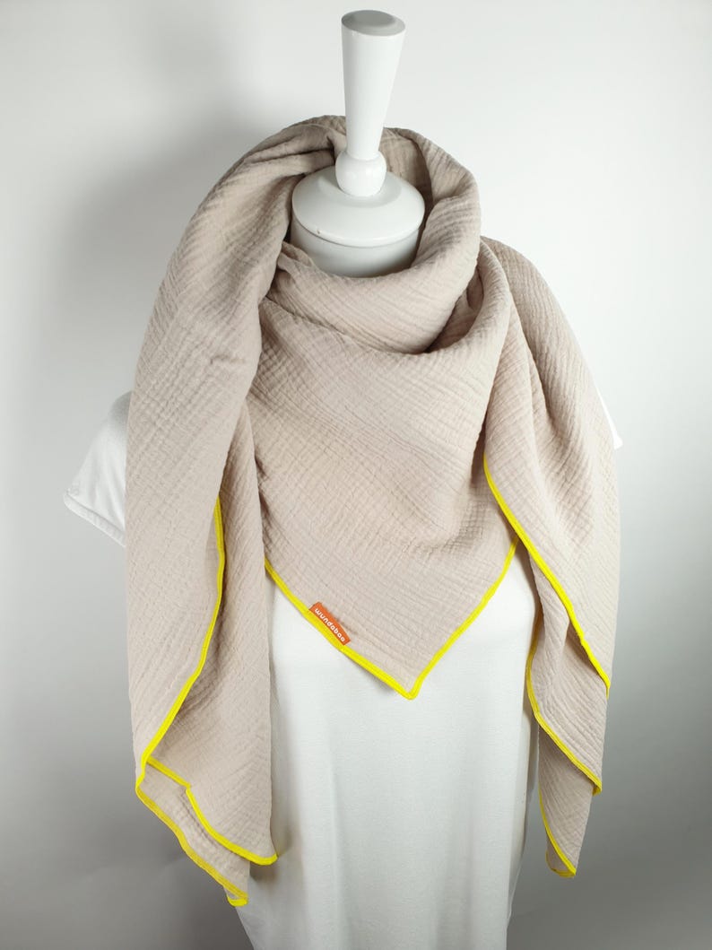 Puede incluir: Una bufanda de algod&oacute;n beige con ribete amarillo. La bufanda est&aacute; doblada y drapeada sobre un maniqu&iacute; blanco.