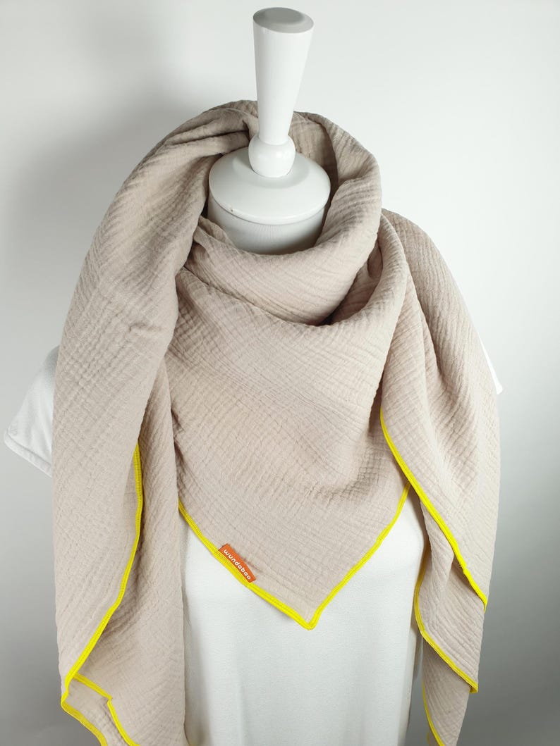 Puede incluir: Una bufanda de algod&oacute;n beige con ribete amarillo. La bufanda est&aacute; doblada y drapeada sobre un maniqu&iacute; blanco. El nombre de la marca "Wunderbar" es visible en la bufanda.