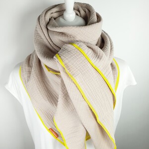 Puede incluir: Una bufanda de algod&oacute;n beige con ribete amarillo. La bufanda est&aacute; doblada y drapeada sobre un maniqu&iacute; blanco.