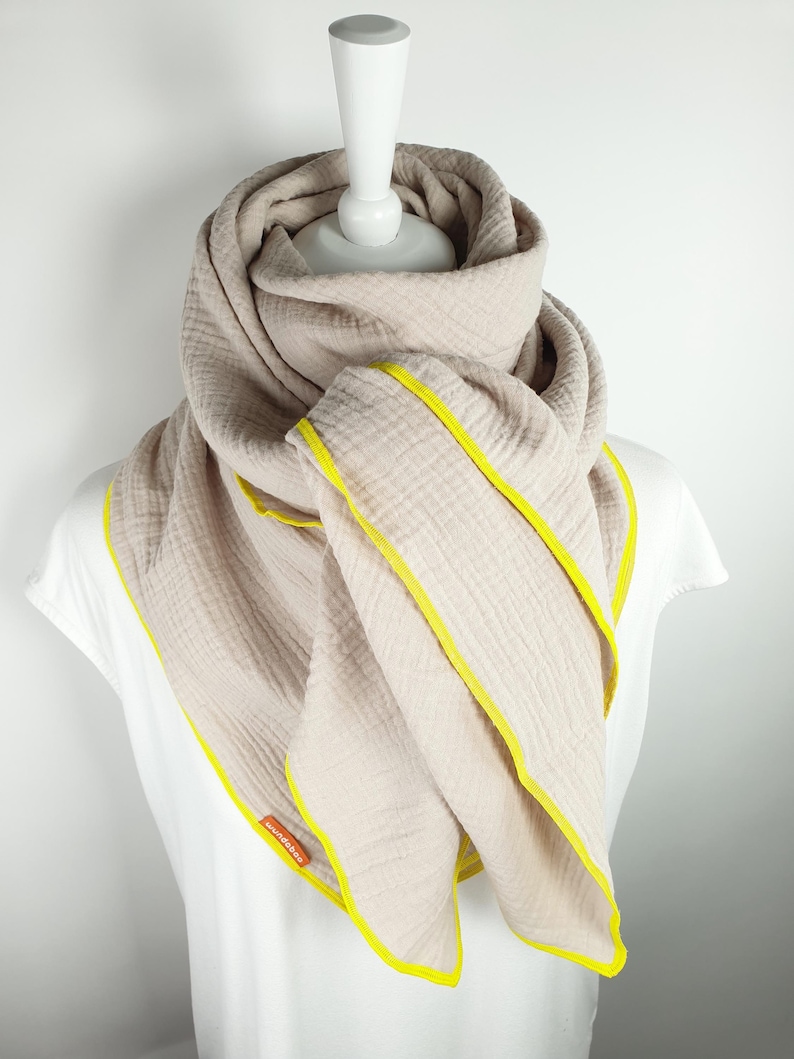 Puede incluir: Una bufanda de algod&oacute;n beige con ribete amarillo. La bufanda est&aacute; doblada y drapeada sobre un maniqu&iacute; blanco.