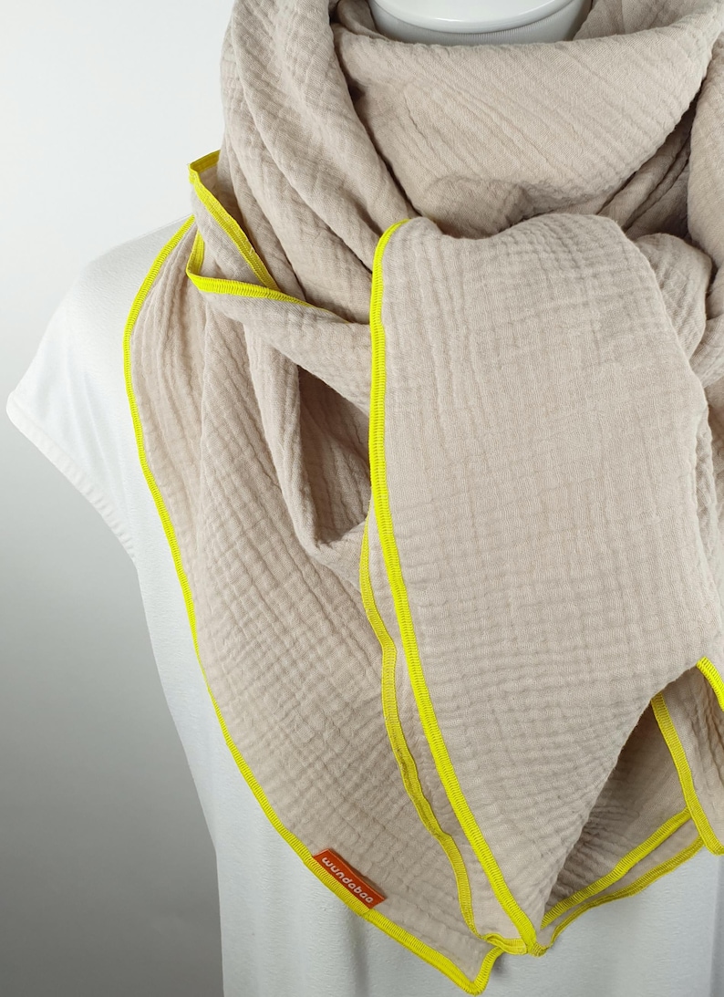 Puede incluir: Una bufanda de algod&oacute;n beige con ribete amarillo. La bufanda est&aacute; doblada y drapeada alrededor del cuello de una persona. El nombre de la marca "wundabar" es visible en la bufanda.